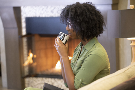 Black African American woman resting at home drinking coffee or CBD tea while relaxing on a couch by a fireplace.  She is holding a warm cup in a living room.の写真素材