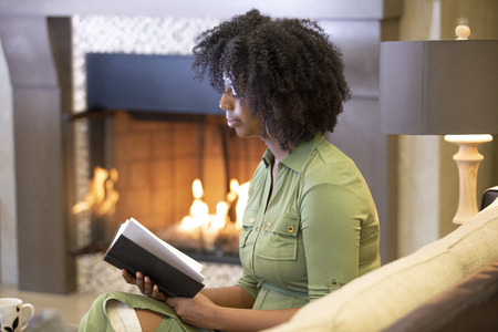 Black African American female resting at home and reading a book in a living room by a fireplace.  The woman is relaxing on a sofa in an apartment with a novel.の写真素材