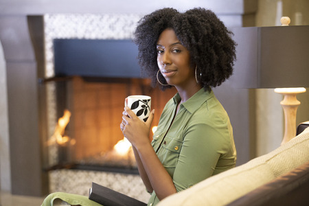 Black African American woman resting at home drinking coffee or CBD tea while relaxing on a couch by a fireplace.  She is holding a warm cup in a living room.の写真素材