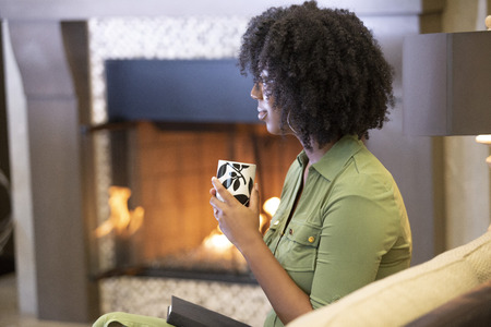 Black African American woman resting at home drinking coffee or CBD tea while relaxing on a couch by a fireplace.  She is holding a warm cup in a living room.の写真素材