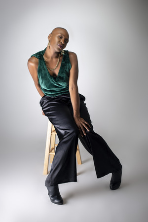Black African American female fashion model wearing trendy green outfit and heels sitting on a stool.  She is confidently showing off a bold bald hairstyle.  の写真素材