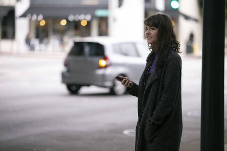 Female pedestrian waiting on a sidewalk for a rideshare.  She is sharing her gps location via cellphone app so the driver can pick her up in the city.  Cars are blurred to obscure make model and license plates. の写真素材