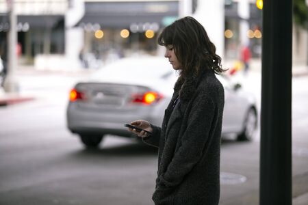 Female pedestrian waiting on a sidewalk for a rideshare.  She is sharing her gps location via cellphone app so the driver can pick her up in the city.  Cars are blurred to obscure make model and license plates. の写真素材