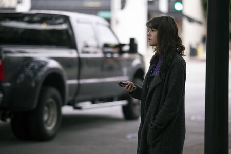 Female pedestrian waiting on a sidewalk for a rideshare.  She is sharing her gps location via cellphone app so the driver can pick her up in the city.  Cars are blurred to obscure make model and license plates.の写真素材