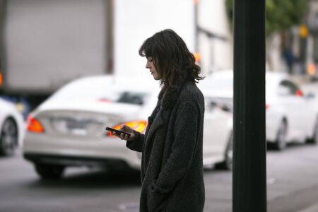 Female pedestrian waiting on a sidewalk for a rideshare.  She is sharing her gps location via cellphone app so the driver can pick her up in the city.  Cars are blurred to obscure make model and license plates.の写真素材