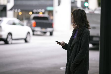Female pedestrian waiting on a sidewalk for a rideshare.  She is sharing her gps location via cellphone app so the driver can pick her up in the city.  Cars are blurred to obscure make model and license plates.の写真素材