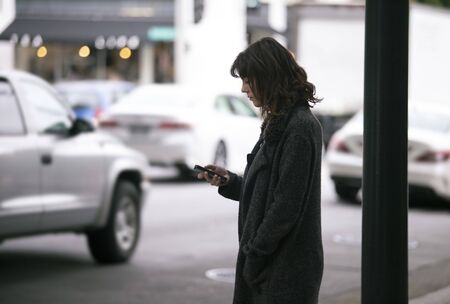 Female pedestrian waiting on a sidewalk for a rideshare.  She is sharing her gps location via cellphone app so the driver can pick her up in the city.  Cars are blurred to obscure make model and license plates.の写真素材