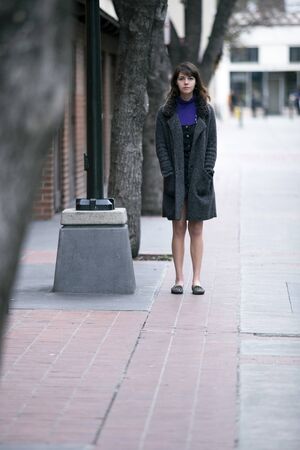 Young pretty woman walking on an urban street sidewalk or a pavement city park.  She looks like an independent millennial female tourist wearing a coat traveling solo on foot.の写真素材