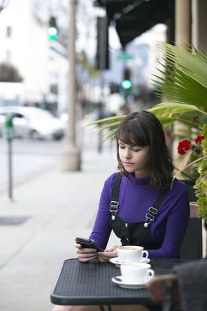 Woman sitting outside a coffee shop or restaurant browsing social media on mobile phone while waiting or hanging out.  She could also be rating or reviewing the food and service of the cafe. の写真素材