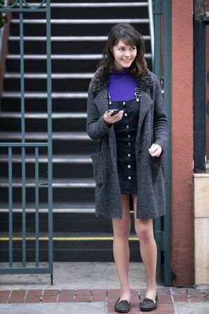 Young female millennial waiting outside apartment home by the stairs for a rideshare or taxi cab.  She is holding her mobile phone and messaging or using app.の写真素材
