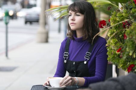 Woman hanging out outdoors and drinking coffee at a side walk cafe or restaurant.  Depicts city life and urban lifestyle.  She is holding a cup while sitting.の写真素材
