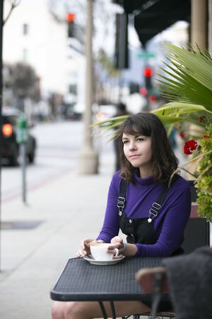 Woman hanging out outdoors and drinking coffee at a side walk cafe or restaurant.  Depicts city life and urban lifestyle.  She is holding a cup while sitting.の写真素材