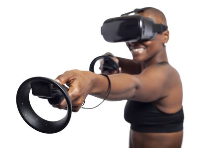 Black female gamer holding virtual reality controllers or wands and wearing a VR headset.  She is playing an interactive video game.  Depicts 3d technology.の写真素材