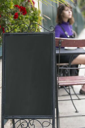 Empy blank sign board in front of a restaurant cafe or coffee shop with a female customer sitting down and waiting.  The image has copy space for logos or text. Depicts advertising and marketing.の写真素材