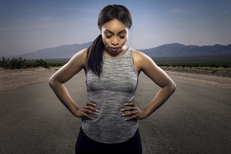 Athletic black African American female runner or jogger resting to pose with a motivated facial expression with a road during sunset in the background.  Depicts endurance and determination during marathon sports.の写真素材