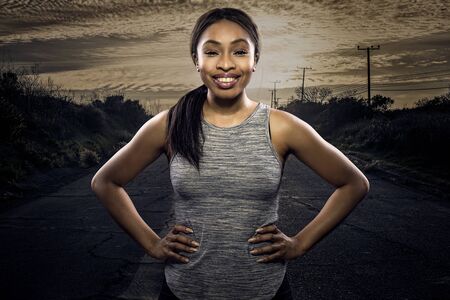 Athletic black African American female runner or jogger resting to pose with a motivated facial expression with a road during sunset in the background.  Depicts endurance and determination during marathon sports.の写真素材