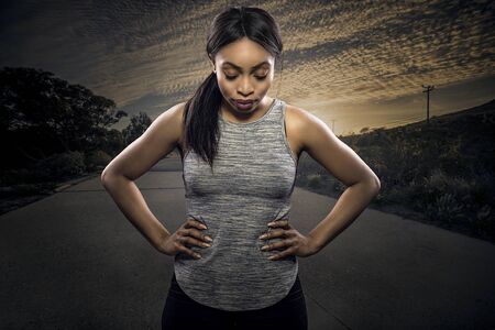 Athletic black African American female runner or jogger resting to pose with a motivated facial expression with a road during sunset in the background.  Depicts endurance and determination during marathon sports.の写真素材
