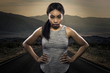 Athletic black African American female runner or jogger resting to pose with a motivated facial expression with a road during sunset in the background.  Depicts endurance and determination during marathon sports.の写真素材