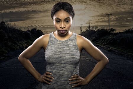 Athletic black African American female runner or jogger resting to pose with a motivated facial expression with a road during sunset in the background.  Depicts endurance and determination during marathon sports.の写真素材