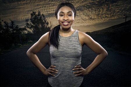 Athletic black African American female runner or jogger resting to pose with a motivated facial expression with a road during sunset in the background.  Depicts endurance and determination during marathon sports.の写真素材