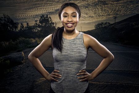 Athletic black African American female runner or jogger resting to pose with a motivated facial expression with a road during sunset in the background.  Depicts endurance and determination during marathon sports.の写真素材