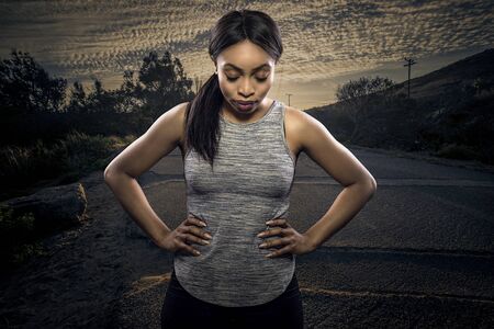 Athletic black African American female runner or jogger resting to pose with a motivated facial expression with a road during sunset in the background.  Depicts endurance and determination during marathon sports.の写真素材