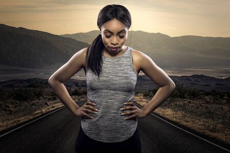Athletic black African American female runner or jogger resting to pose with a motivated facial expression with a road during sunset in the background.  Depicts endurance and determination during marathon sports.の写真素材