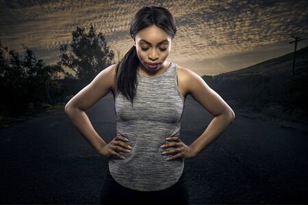 Athletic black African American female runner or jogger resting to pose with a motivated facial expression with a road during sunset in the background.  Depicts endurance and determination during marathon sports.の写真素材