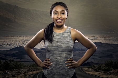 Athletic black African American female runner or jogger resting to pose with a motivated facial expression with a road during sunset in the background.  Depicts endurance and determination during marathon sports.の写真素材