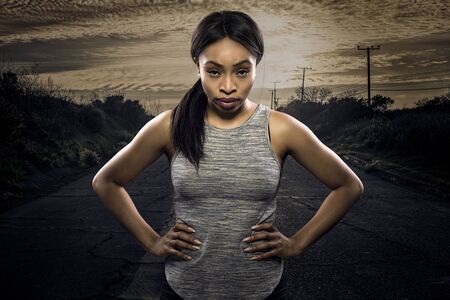 Athletic black African American female runner or jogger resting to pose with a motivated facial expression with a road during sunset in the background.  Depicts endurance and determination during marathon sports.の写真素材