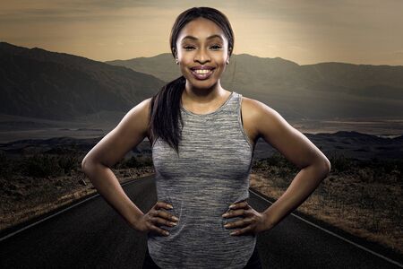 Athletic black African American female runner or jogger resting to pose with a motivated facial expression with a road during sunset in the background.  Depicts endurance and determination during marathon sports.の写真素材