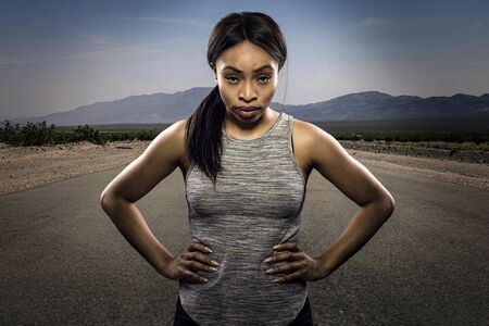 Athletic black African American female runner or jogger resting to pose with a motivated facial expression with a road during sunset in the background.  Depicts endurance and determination during marathon sports.の写真素材