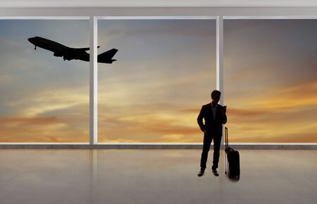 Silhouette of a traveling businessman with luggage in front of an airport window with a view of the sky and an airplane.  The panorama has copy space for text.  Depicts business travel.の写真素材