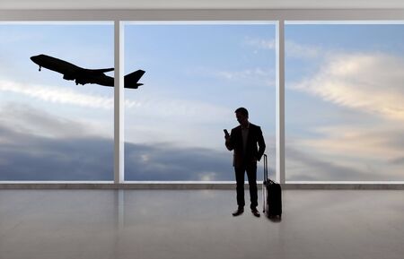Silhouette of a traveling businessman with luggage in front of an airport window with a view of the sky and an airplane.  The panorama has copy space for text.  Depicts business travel.の写真素材