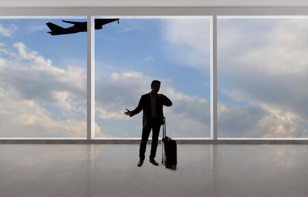 Silhouette of a traveling businessman with luggage in front of an airport window with a view of the sky and an airplane.  The panorama has copy space for text.  Depicts business travel.の写真素材