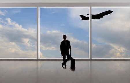 Silhouette of a traveling businessman with luggage in front of an airport window with a view of the sky and an airplane.  The panorama has copy space for text.  Depicts business travel.の写真素材