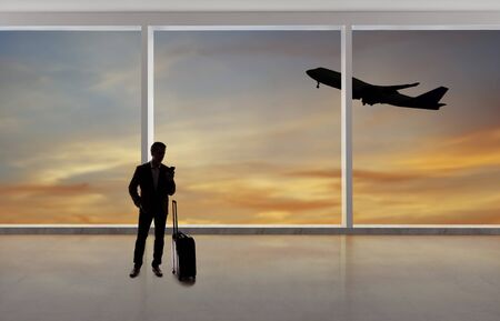 Silhouette of a traveling businessman with luggage in front of an airport window with a view of the sky and an airplane.  The panorama has copy space for text.  Depicts business travel.の写真素材