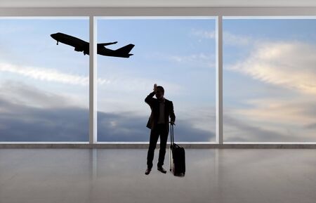Silhouette of a traveling businessman with luggage in front of an airport window with a view of the sky and an airplane.  The panorama has copy space for text.  Depicts business travel.の写真素材