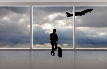 Silhouette of a traveling businessman with luggage in front of an airport window with a view of the sky and an airplane.  The panorama has copy space for text.  Depicts business travel.の写真素材