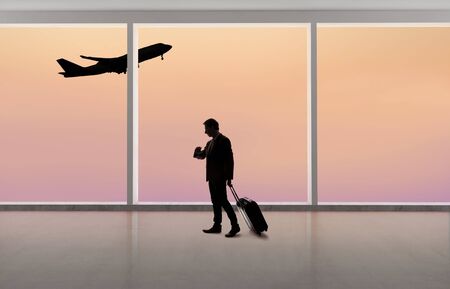 Silhouette of a traveling businessman with luggage in front of an airport window with a view of the sky and an airplane.  The panorama has copy space for text.  Depicts business travel.の写真素材