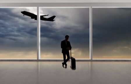 Silhouette of a traveling businessman with luggage in front of an airport window with a view of the sky and an airplane.  The panorama has copy space for text.  Depicts business travel.の写真素材