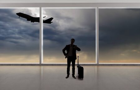 Silhouette of a traveling businessman with luggage in front of an airport window with a view of the sky and an airplane.  The panorama has copy space for text.  Depicts business travel.の写真素材