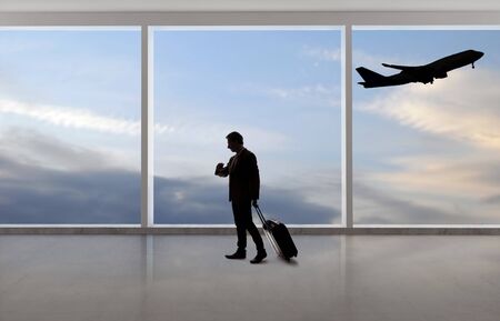 Silhouette of a traveling businessman with luggage in front of an airport window with a view of the sky and an airplane.  The panorama has copy space for text.  Depicts business travel.の写真素材