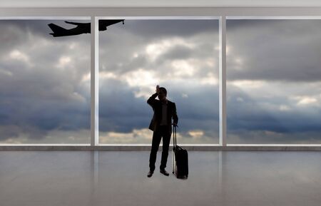 Silhouette of a traveling businessman with luggage in front of an airport window with a view of the sky and an airplane.  The panorama has copy space for text.  Depicts business travel.の写真素材