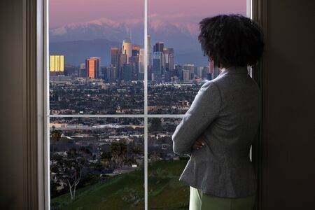 Black female business woman looking out the window of an office in Los Angeles.  She looks like a female architect thinking of urban development or a city mayor or governor planning zoning laws. の写真素材