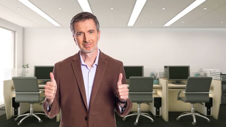 Middle-Aged businessman with gray hair and wearing a brown jacket standing in an office.  Depicts startup and corporate business.  He is holding thumbs up in approval.の写真素材