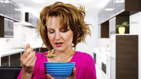 Caucasian female with frizzy hair sitting on a couch eating a breakfast bowl of cereal.  She is at home and looks like she just woke up and doing a morning routine. の写真素材