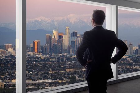 Businessman wearing a suit looking at the buildings of downtown Los Angeles from an office window.  The man looks like a politician like a mayor, or an architect or a real estate developer working in LA.の写真素材