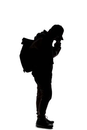 Silhouette of a man wearing a backpack looking like a traveler or hiker trekking. He is using a cell phone の写真素材
