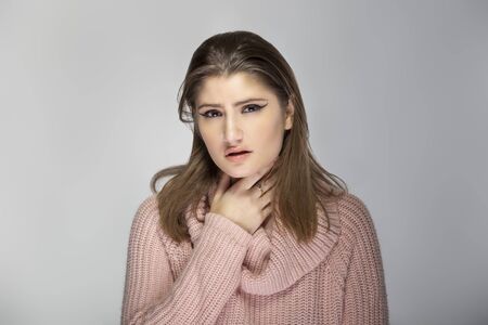 Close up portrait of a young Caucasian woman wearing a pink sweater on a grey background.  The model looks sick with a sore throatの写真素材